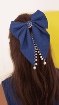 A large denim bow hair accessory with dangling pearl bead strands, shown worn on a girl’s brown hair from the back.