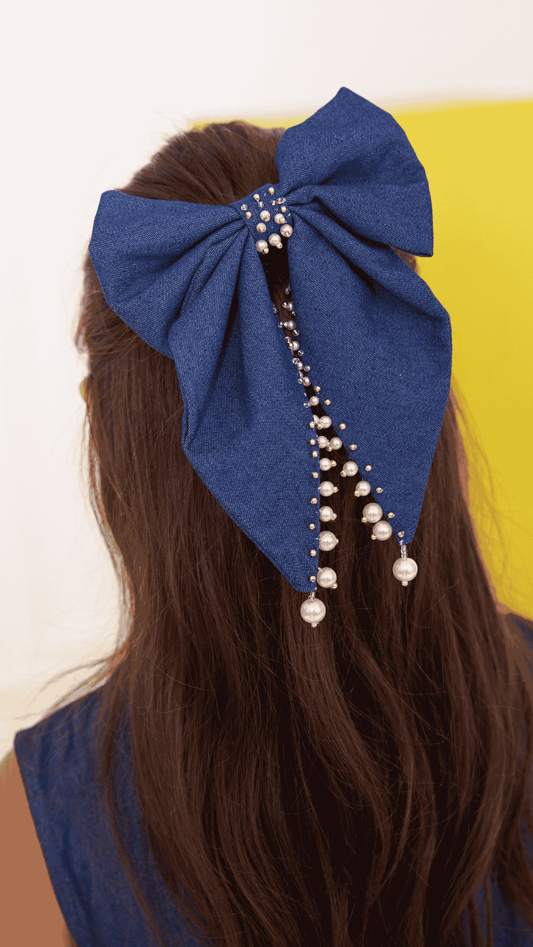 A large denim bow hair accessory with dangling pearl bead strands, shown worn on a girl’s brown hair from the back.