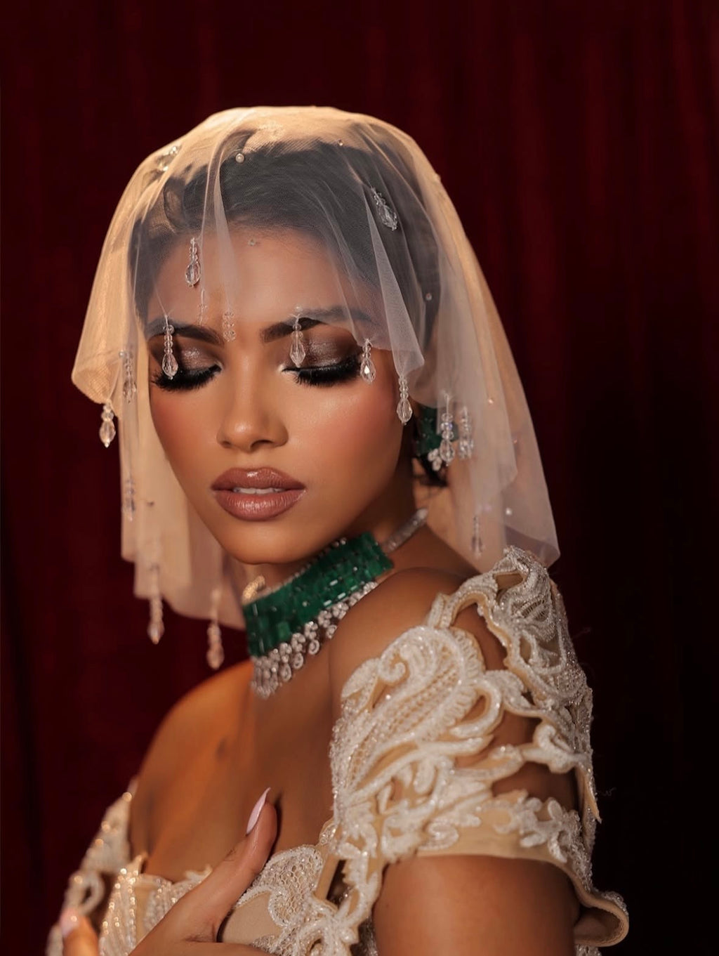 A woman wearing an ornate cream lace bridal gown with a sheer embroidered veil, a green beaded choker, and dangling earrings, posed against a dark red curtain background.