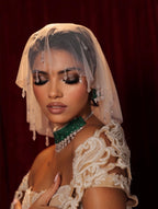 A woman wearing an ornate cream lace bridal gown with a sheer embroidered veil, a green beaded choker, and dangling earrings, posed against a dark red curtain background.