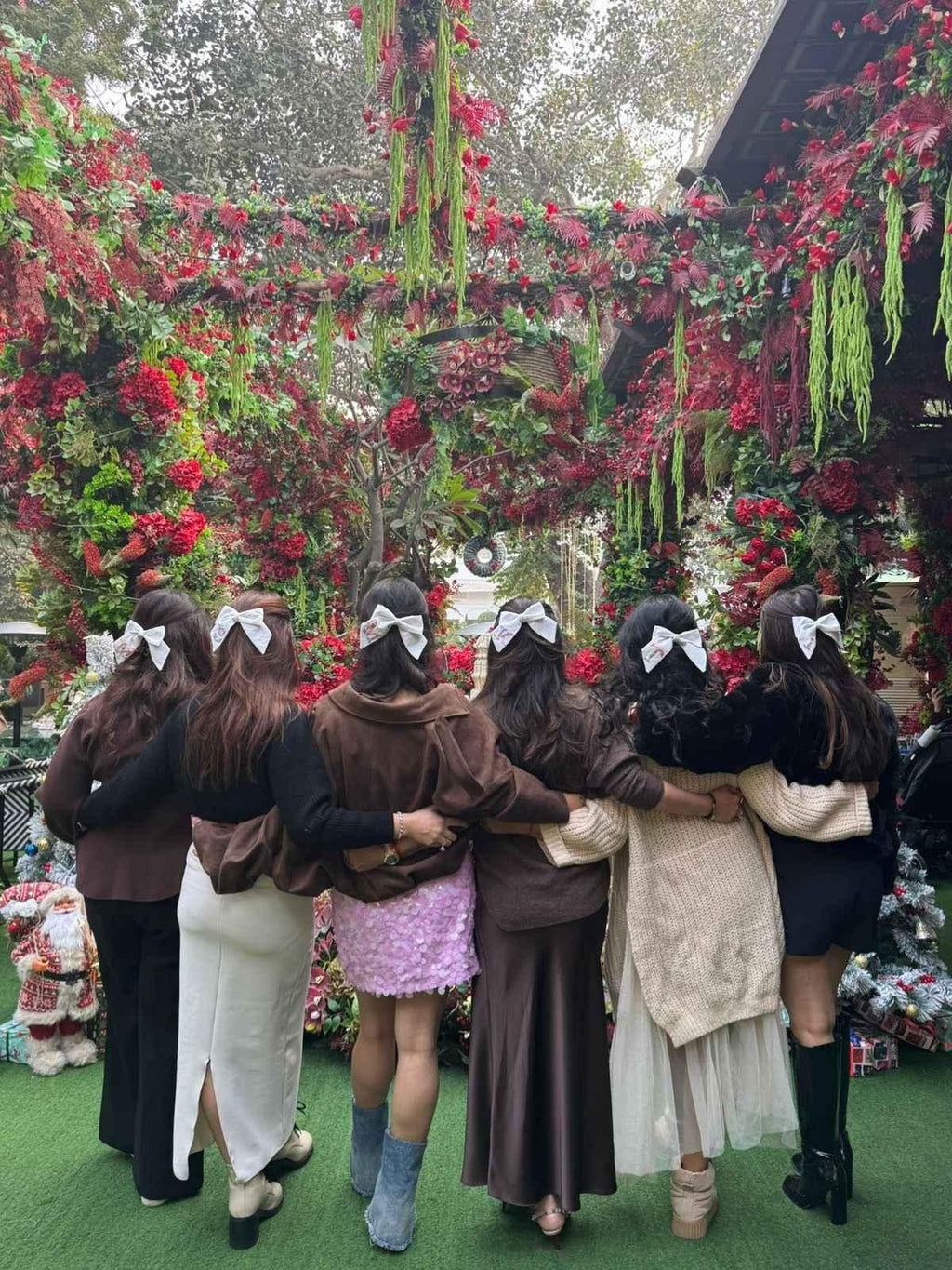Group of women wearing handmade hair bows, posing outdoors amid lush floral decorations.