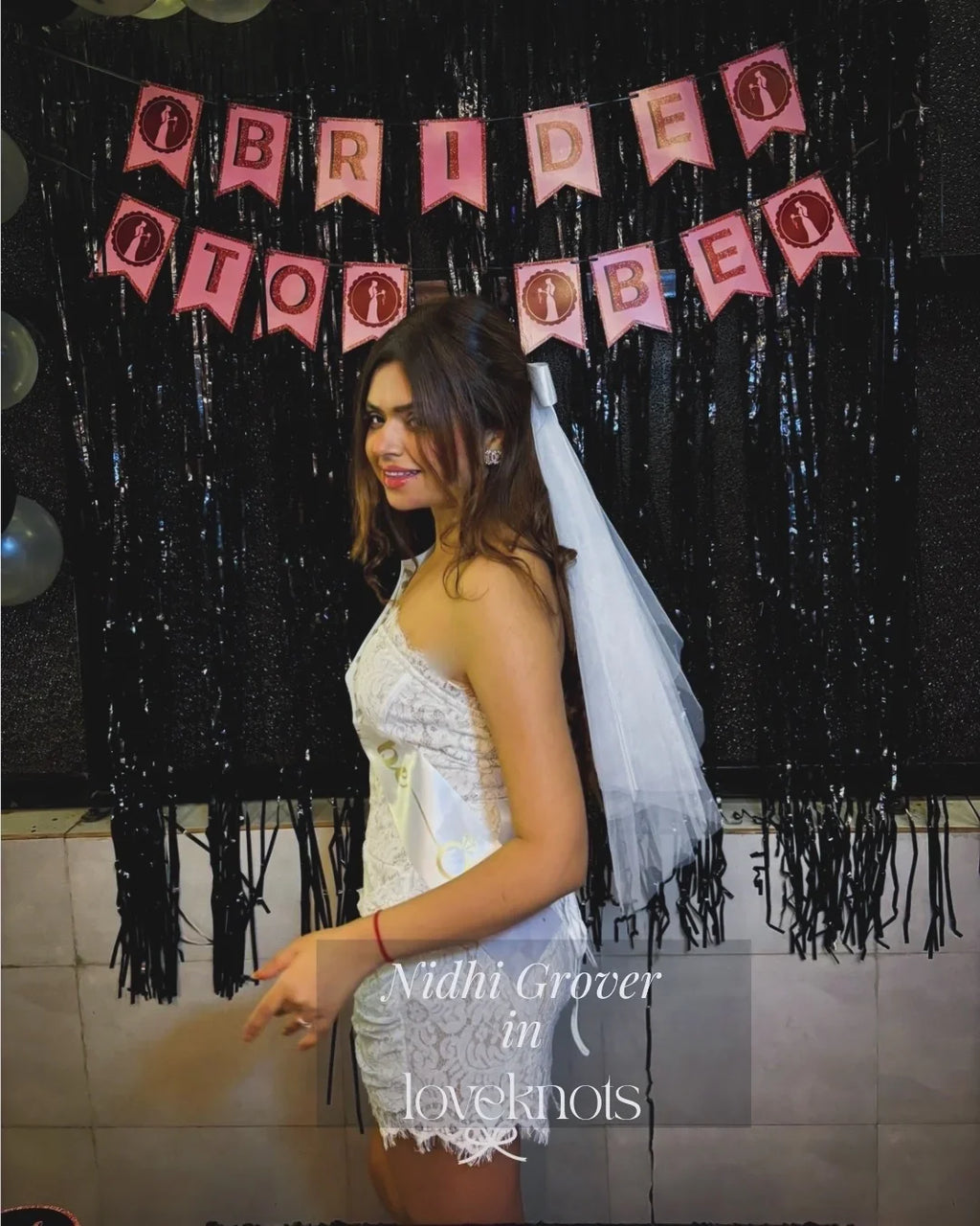 A woman in a white lace bridal-style dress with a veil stands in front of a black tinsel backdrop and pink 'Bride to Be' banner. The photo includes a visible watermark reading 'Nidhi Grover in loveknots'.