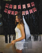 A woman in a white lace bridal-style dress with a veil stands in front of a black tinsel backdrop and pink 'Bride to Be' banner. The photo includes a visible watermark reading 'Nidhi Grover in loveknots'.