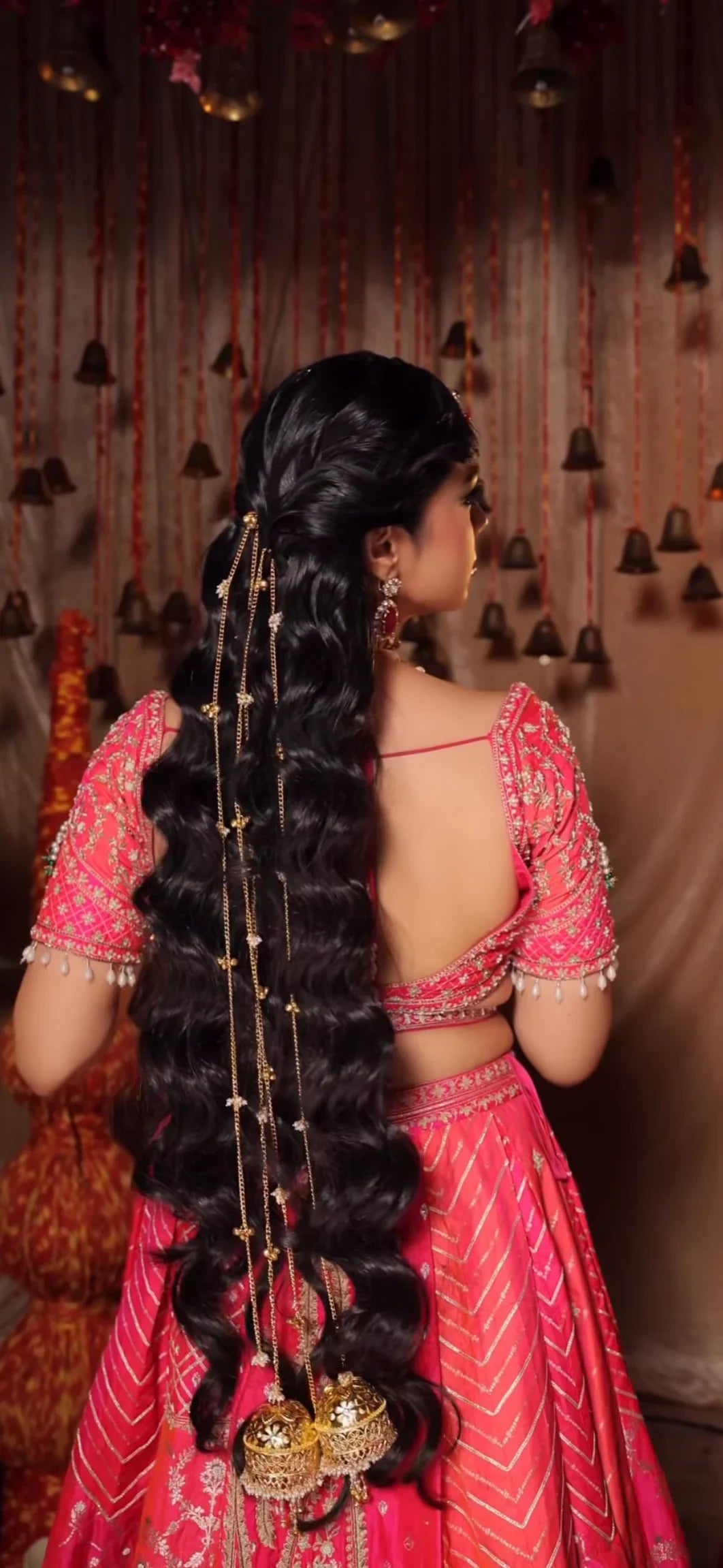 A woman shown from the back wearing a bright pink traditional outfit with long, elaborate black hair adorned with gold plated hair chain and jhumki , set against a decorated indoor backdrop with hanging bells.