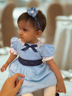Baby in a blue dress with a plumeria flower headband, sitting on a chair.
