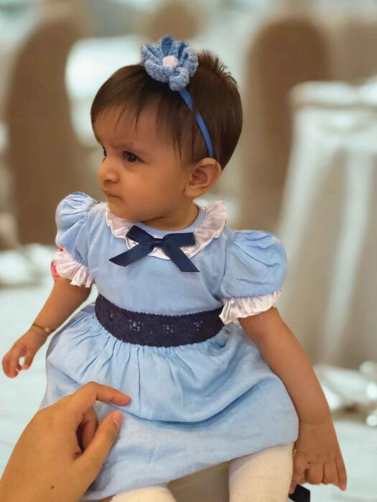 Baby in a blue dress with a plumeria flower headband, sitting on a chair.