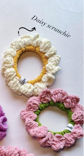 Two handmade crocheted scrunchies on a white background, one cream and yellow and labeled 'Daisy scrunchie', and one pink and green. A partial purple scrunchie is visible at the bottom left.