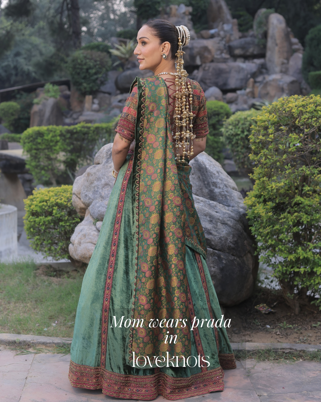 A woman stands outdoors in a garden wearing a traditional green embroidered dress with a matching dupatta and ornate gold jewelry. The text overlay reads 'Mom wears prada in loveknots'.