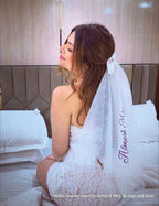 Woman wearing a white bridal veil with 'Almost Mrs.' text, sitting on a bed.