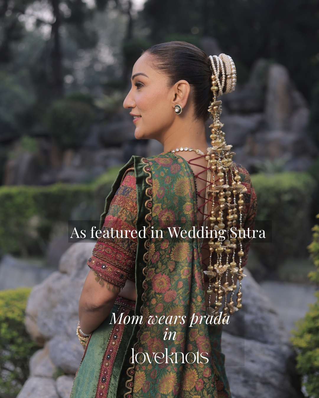 A woman photographed from the back in a garden setting, wearing a green patterned traditional Indian sari with ornate gold jewelry and a long decorative hair accessory. White text overlays read 'As featured in Wedding Snra' and 'Mom wears prada in loveknos'.