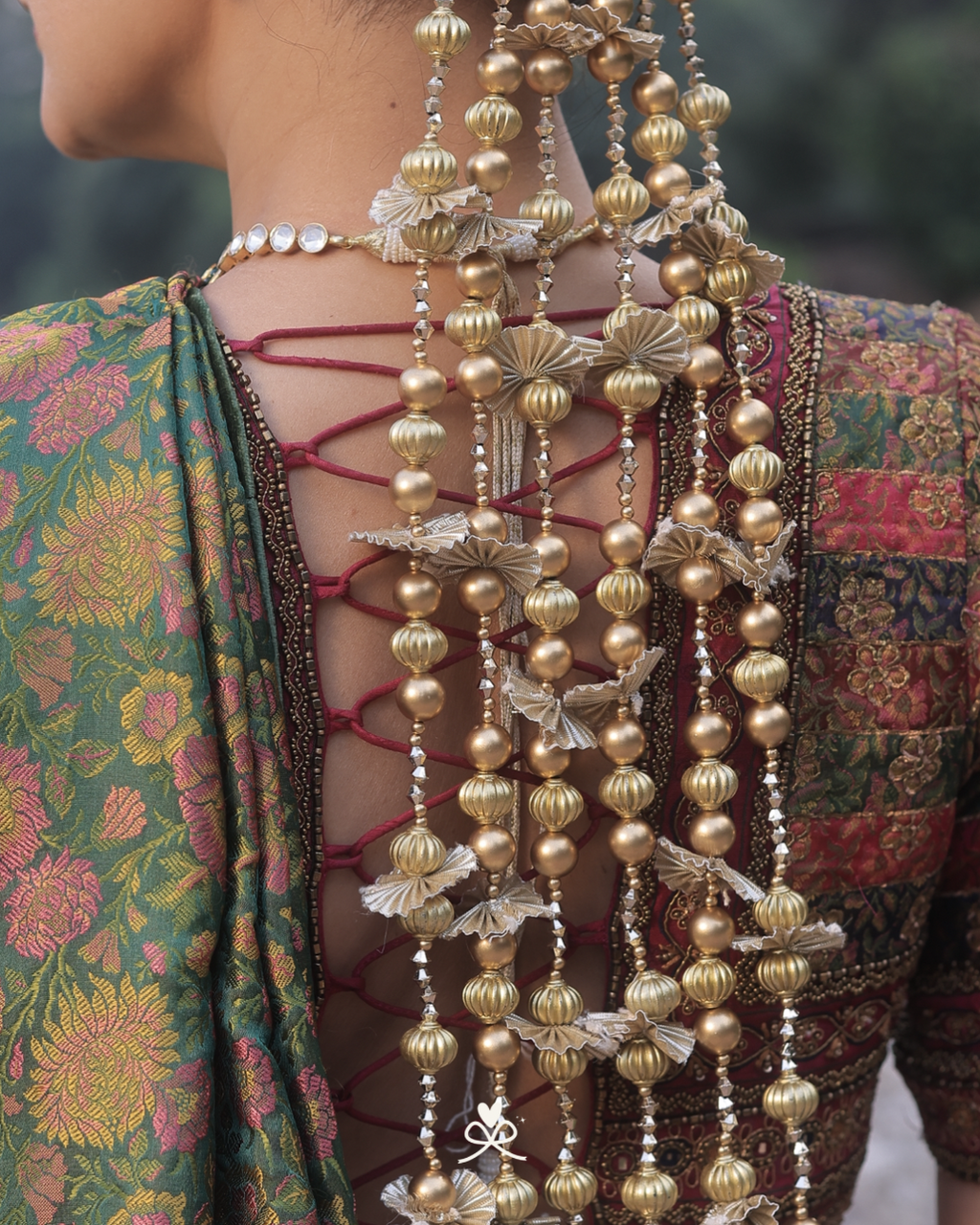 Close-up of a person's back wearing an ornate traditional outfit with a green floral patterned fabric, lace-up open back, and multiple strands of gold and silver beaded hair ornaments.