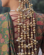 Close-up of a person's back wearing an ornate traditional outfit with a green floral patterned fabric, lace-up open back, and multiple strands of gold and silver beaded hair ornaments.