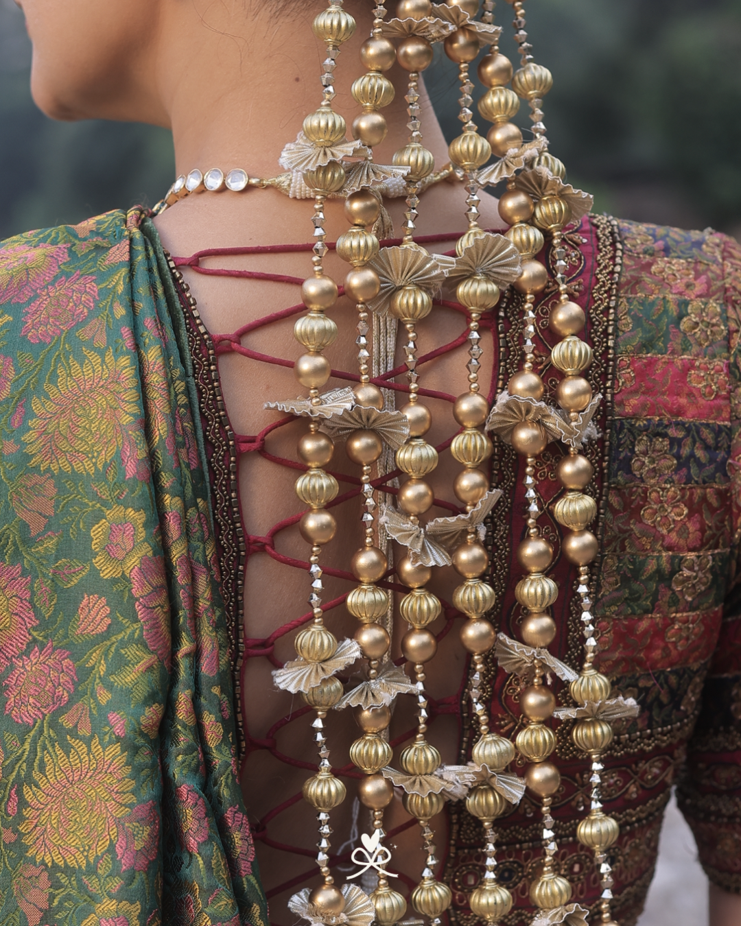 Close-up of a person's back wearing an ornate traditional outfit with a green floral patterned fabric, lace-up open back, and multiple strands of gold and silver beaded hair ornaments.