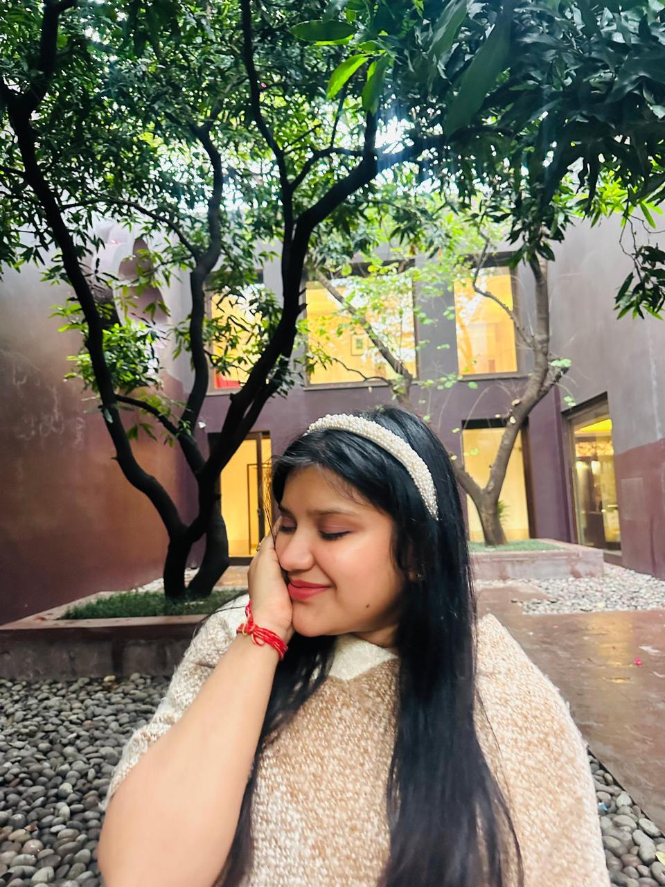 Woman with long dark hair and a pearl handmandeheadband sitting outdoors with trees and building in the background