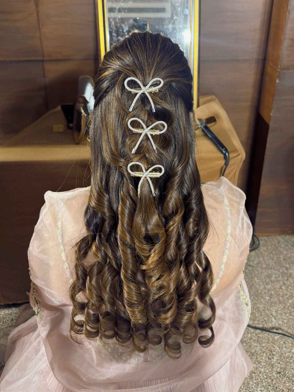 Person with styled hair featuring bows, wearing a light-colored dress, in front of a mirror.